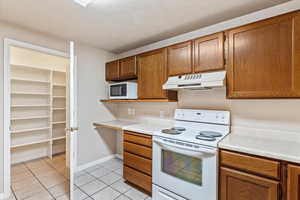 Kitchen featuring under cabinet range hood, white appliances, light tile patterned floors, light countertops, and brown cabinetry