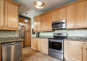 Kitchen with appliances with stainless steel finishes, light brown cabinets, stone countertops, hanging light fixtures, and light tile patterned floors