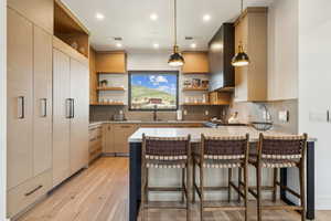Kitchen with open shelves, a sink, a peninsula, wall chimney range hood, and light wood-type flooring