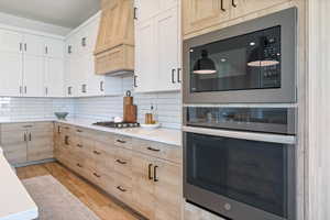 Kitchen with appliances with stainless steel finishes, custom exhaust hood, decorative backsplash, light brown cabinets, and light wood finished floors