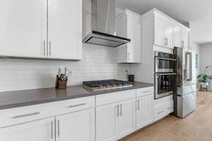Kitchen with wall chimney range hood, stainless steel appliances, white cabinetry, dark countertops, and light wood-style floors