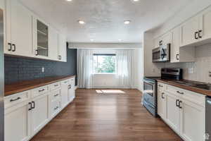 Kitchen with wooden counters, appliances with stainless steel finishes, glass insert cabinets, backsplash, and dark wood finished floors