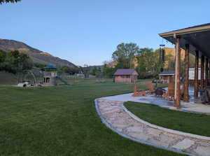 View of green lawn featuring a playground, a patio, a mountain view, an outbuilding, and an outdoor fire pit