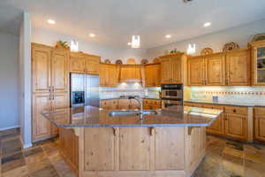 Kitchen featuring appliances with stainless steel finishes, a sink, a kitchen island with sink, stone tile flooring, and decorative backsplash