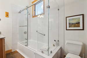 Bathroom with combined bath / shower with glass door, toilet, vanity, baseboards, and stone finish flooring
