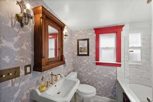 Bathroom with toilet, a sink, baseboards, wallpapered walls, and a bath