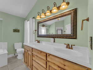 Bathroom with toilet, double vanity, vaulted ceiling, tile patterned flooring, and a tub to relax in