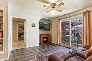 Living area featuring a textured ceiling, a warm lit fireplace, wood finished floors, ceiling fan, and built in features