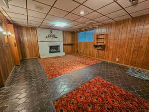 Unfurnished living room with wood walls, a fireplace, a paneled ceiling, and baseboards