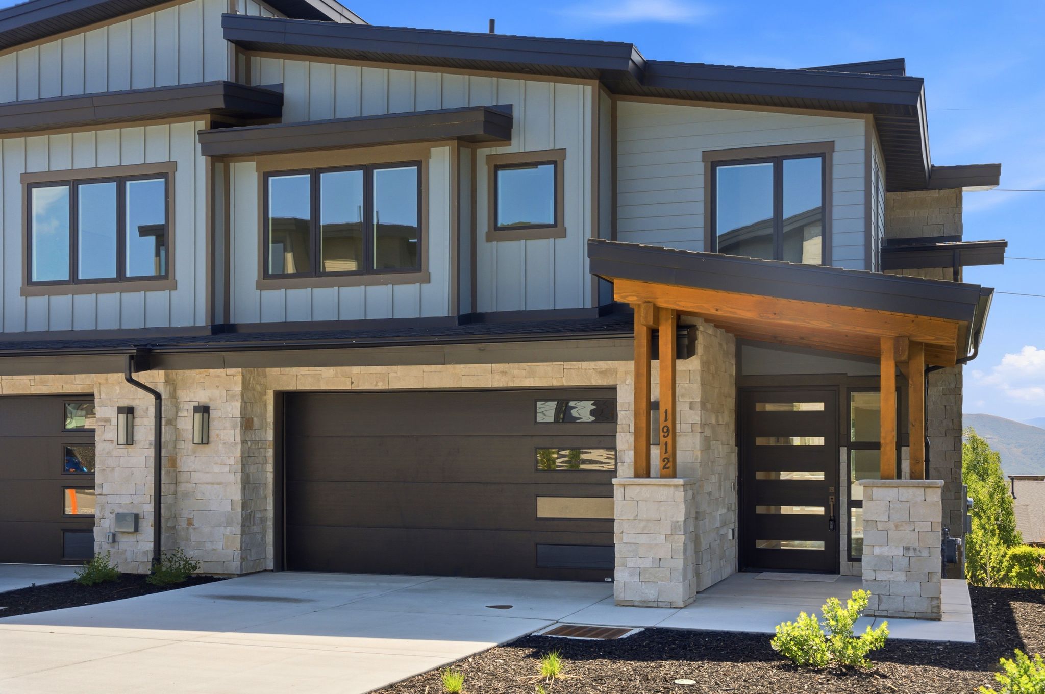 Contemporary exterior featuring vertical and horizontal siding, stone facade accents, an angled wood-beam entry canopy, modern dark-framed windows, and a dark garage door with horizontal frosted glass inserts