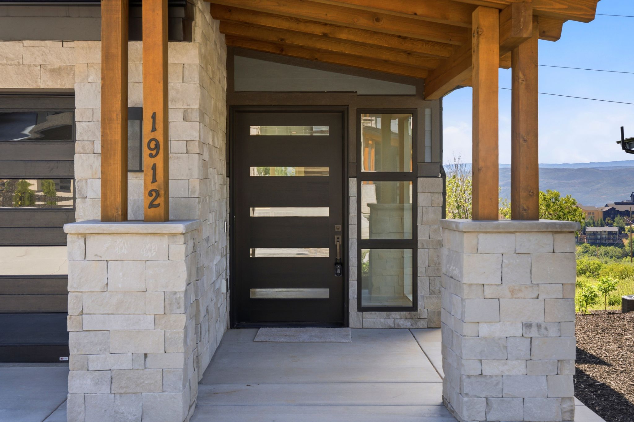 Contemporary entry featuring a dark-finish door with horizontal lites, light-toned stone masonry, and exposed wood beams