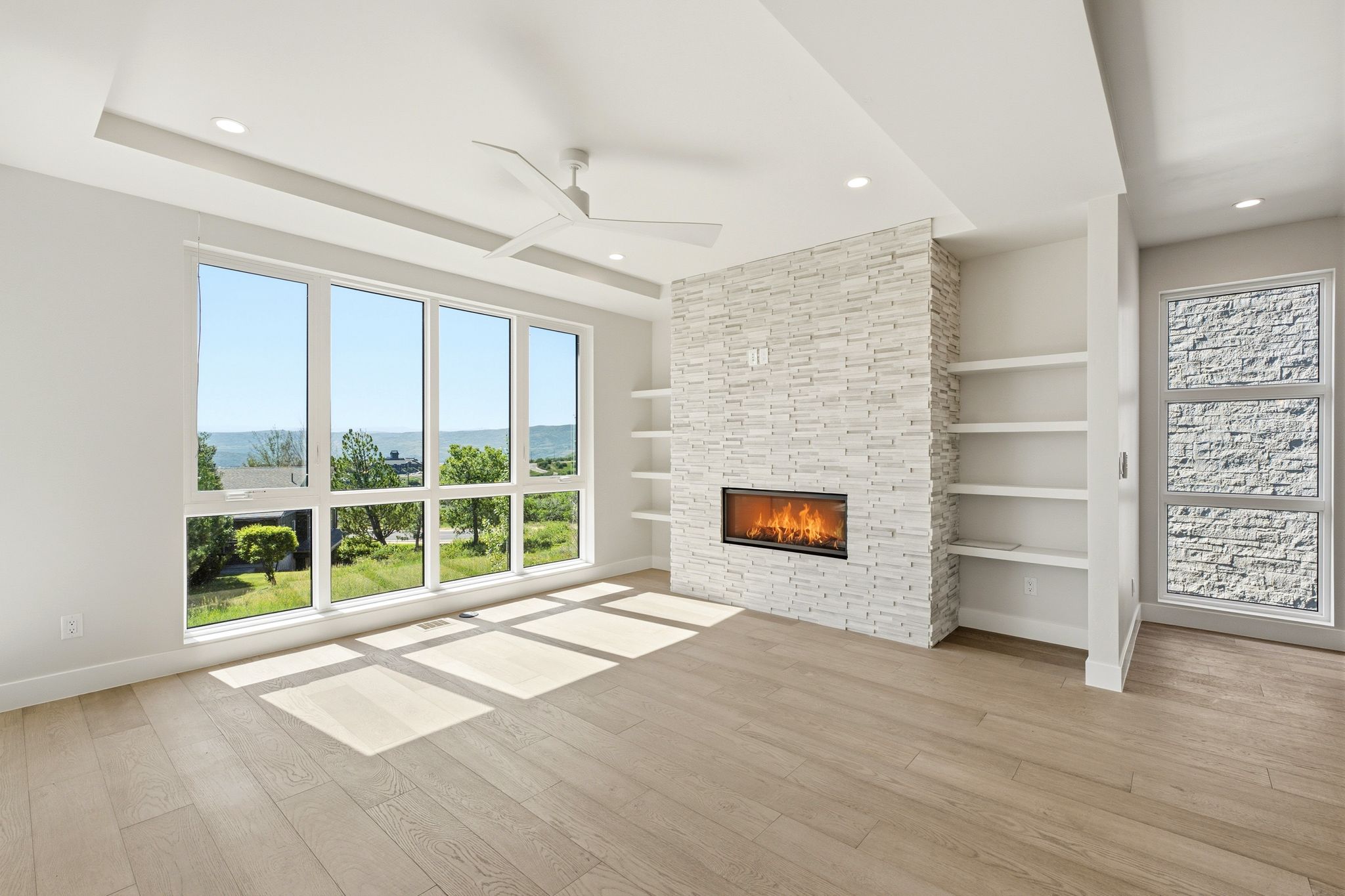Spacious room featuring expansive windows, wood-finish flooring, a contemporary fireplace with a stacked stone surround, built-in shelving, and a ceiling fan