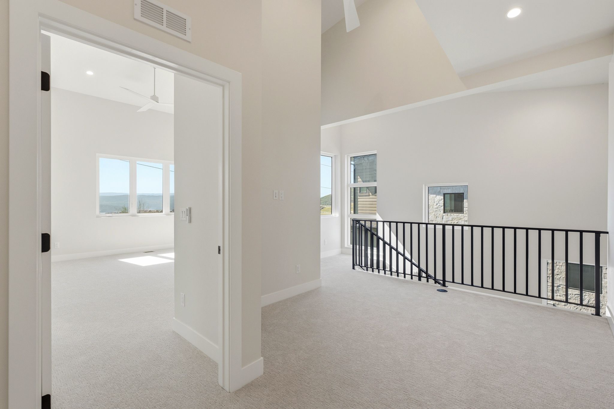 Carpeted hallway with black metal railing, leading to a room with multiple windows overlooking a body of water