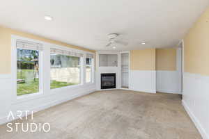 Unfurnished living room featuring a ceiling fan, a wainscoted wall, carpet, healthy amount of natural light, and a glass covered fireplace