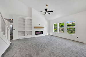Unfurnished living room featuring high vaulted ceiling, a glass covered fireplace, a ceiling fan, stairway, and carpet floors