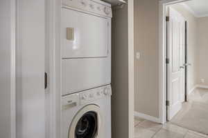 Washroom featuring stacked washer and clothes dryer, ornamental molding, baseboards, light tile patterned floors, and light colored carpet