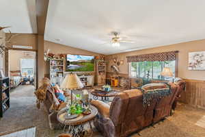 Living room with carpet, ceiling fan, a wainscoted wall, wood walls, and lofted ceiling