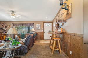 Living room featuring a wainscoted wall, wood walls, carpet, ceiling fan, and a dry bar