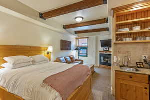 Bedroom with carpet floors, beamed ceiling, bar with sink, and a glass covered fireplace