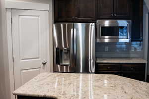 Kitchen with appliances with stainless steel finishes, backsplash, dark brown cabinetry, and light stone counters