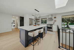 Kitchen featuring open shelves, appliances with stainless steel finishes, wall chimney exhaust hood, beverage cooler, and light wood finished floors