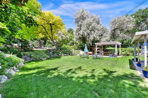 View of green lawn featuring a pergola, a patio, and view of scattered trees in a terraced yard with amply perennial flowers and water feature