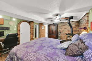 Carpeted bedroom featuring brick wall, a wood stove, a textured ceiling, ceiling fan, and an office area
