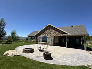 Rear view of house featuring a fire pit, a patio, a yard, stone siding, and roof with shingles