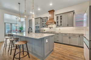 Kitchen with gray cabinetry, custom exhaust hood, appliances with stainless steel finishes, a center island with sink, and a kitchen bar