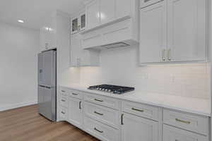 Kitchen with stainless steel appliances, white cabinetry, glass insert cabinets, light wood-type flooring, and decorative backsplash