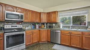 Kitchen featuring appliances with stainless steel finishes, a sink, decorative backsplash, brown cabinetry, and dark countertops