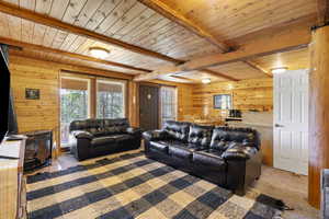 Living room with a wood stove, wooden walls, carpet floors, and wooden ceiling with exposed beams