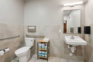 Half bath with toilet, tile patterned flooring, tile walls, wainscoting, and a sink