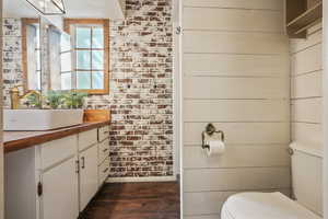 Half bath with wood finished floors, toilet, vanity, baseboards, and brick wall