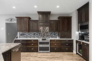 Kitchen featuring appliances with stainless steel finishes, decorative backsplash, dark brown cabinetry, light wood-style flooring, vented hood, and recessed lighting