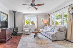 Living area featuring dark wood-style flooring, plenty of natural light, ornamental molding, a wood stove, and a ceiling fan