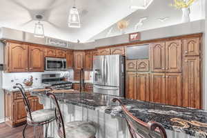 Kitchen featuring appliances with stainless steel finishes, lofted ceiling, brown cabinetry, dark wood-type flooring, and pendant lighting
