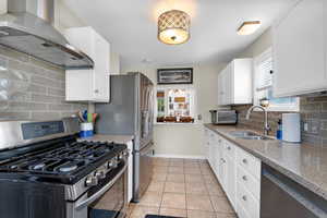 Kitchen with stainless steel appliances, wall chimney range hood, a sink, light stone countertops, and white cabinetry