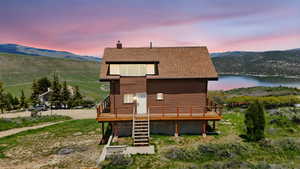 Back of house at dusk with a deck with water view, a chimney, roof with shingles, and stairway