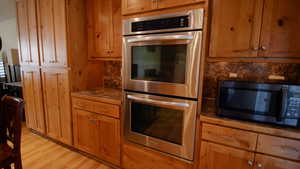 Kitchen with stainless steel double oven, light wood-style flooring, brown cabinets, and dark countertops