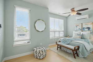 Bedroom with a wall mounted AC, wood finished floors, baseboards, and a ceiling fan