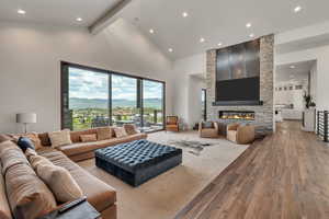 Living area featuring light wood finished floors, high vaulted ceiling, a stone fireplace, recessed lighting, and beam ceiling