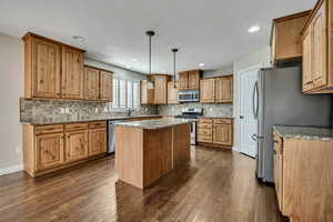 Kitchen with stainless steel appliances, decorative backsplash, dark wood-style flooring, a kitchen island, and light stone countertops