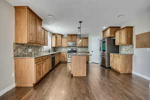 Kitchen with appliances with stainless steel finishes, a kitchen island, baseboards, dark wood-style flooring, and recessed lighting