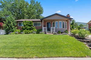 Single story home with a front lawn, covered porch, a chimney, and brick siding