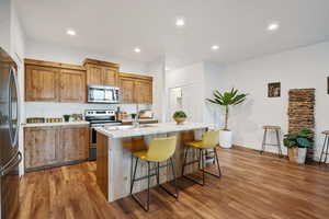 Kitchen featuring appliances with stainless steel finishes, brown cabinetry, a kitchen bar, wood finished floors, and recessed lighting