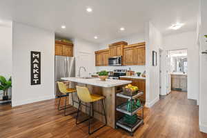 Kitchen featuring appliances with stainless steel finishes, light countertops, brown cabinets, an island with sink, and dark wood-type flooring