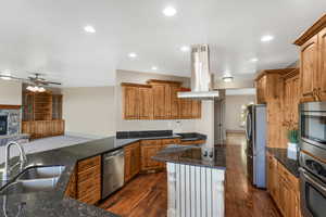 Kitchen with island exhaust hood, appliances with stainless steel finishes, a sink, a ceiling fan, and dark stone countertops