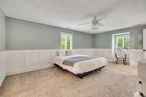 Carpeted bedroom featuring multiple windows, a wainscoted wall, a decorative wall, and ceiling fan