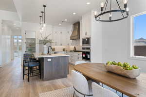 Kitchen with stainless steel appliances, custom range hood, a chandelier, an island with sink, and light wood finished floors